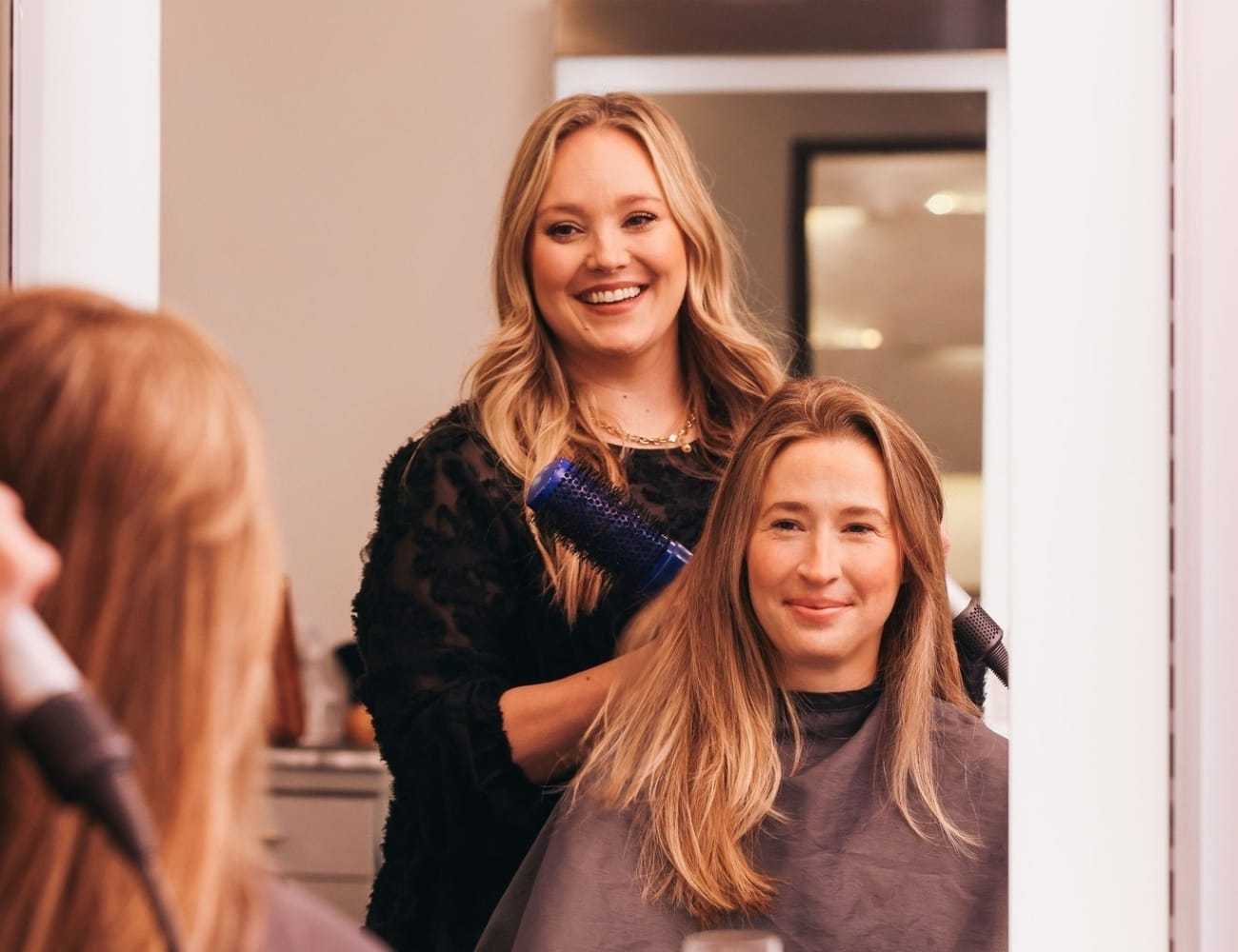 Hairstylist smiling while blow-drying a client's hair in a salon.